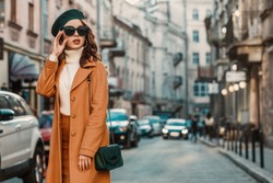 Outdoor autumn portrait of young elegant fashionable woman wearing trendy sunglasses, camel color coat, turtleneck, with textured leather shoulder bag, walking in street of European city. Copy space