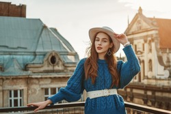 Outdoor fashion portrait of elegant fashionable  woman, model wearing trendy blue knitted sweater, belt, stylish white hat, earrings, posing at sunset, in European city. Copy, empty space for text