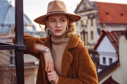 Outdoor fashion portrait of elegant woman wearing beige hat, wrist watch, turtleneck, brown faux fur coat, trendy big earrings with rhinestones, posing in European city. Copy, empty space for text