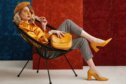 Full-length studio fashion portrait of elegant woman wearing yellow color sunglasses, beret, silk blouse, houndstooth printed trousers, pointed toe shoes, posing on chair, holding stylish leather bag
