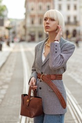 Street fashion photo of elegant  woman wearing trendy checkered blazer, wide wicker belt, holding brown faux croco leather textured bag. Model walking in street of European city
