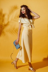 Summer fashion concept: woman wearing monochrome yellow outfit, holding small blue bag, posing in studio on yellow background.