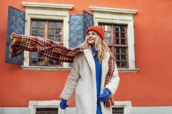 Christmas, winter holidays, lifestyle conception: happy smiling woman wearing white faux fur coat, colorful scarf, hat, walking in street of European city. Copy, empty space for text