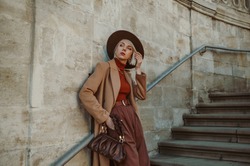 Outdoor autumn fashion portrait of elegant, luxury woman wearing trendy midi beige, camel color coat, brown hat, trousers, orange turtleneck, holding leather pouch bag, posing in street of city