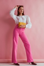 Elegant fashionable woman wearing white vintage blouse with lace collar, pink jeans, holding small padded yellow leather bag, posing on pink background. Full length portrait
