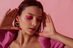 Close up beauty portrait of young beautiful woman with pink, fuchsia color eyeshadow makeup, flawless clean skin, wearing elegant golden earrings, pink blouse. Spring, summer fashion trend