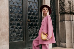 Street style, fashion conception: elegant woman wearing trendy pink suit, burgundy color hat, holding yellow bag, posing in street of city. Copy, empty space for text