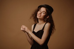 Beautiful happy smiling fashionable curly brunette woman wearing trendy leather beret, golden earrings, stylish black dress, posing in studio on brown background. Copy, empty space for text