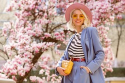 Spring fashion, street style portrait of elegant happy smiling woman wearing pink hat, yellow sunglasses, blue suit, striped t-shirt, holding trendy leather bag, posing outdoor. Copy, empty space