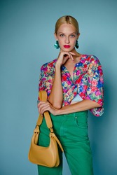 Summer fashion portrait of confident blonde woman wearing colorful blouse, green earrings, high waist jeans, holding trendy yellow leather bag, posing on blue background