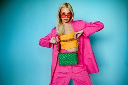 Fashion portrait of confident woman wearing trendy summer pink fuchsia color suit, orange sunglasses, holding green quilted faux leather bag, posing in studio, on blue background. Copy, empty space
