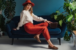 Autumn fashion conception: elegant lady wearing orange hat, vintage style blouse, trendy culottes, leopard print loafer shoes, posing on sofa, in blue interior. Copy, empty space for text