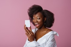 Yong beautiful happy smiling surprised African American woman, model wearing elegant jewelry, classic shirt, holding small white box, posing in studio, on pink background. Copy, empty space for text