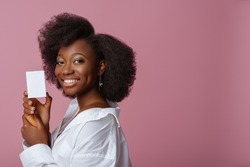 Yong beautiful happy smiling African American woman, model wearing elegant jewelry, classic shirt, holding small white box, posing in studio, on pink background. Copy, empty space for text