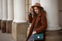 Happy smiling elegant fashionable woman wearing trendy brown faux fur teddy bear coat, with green textured leather shoulder bag, posing in street of European city. Copy, empty space for text