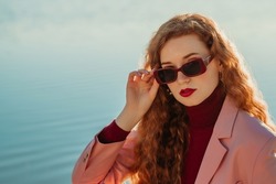 Beautiful freckled readhead woman with natural long curly hair, bold marsala color lips makeup, wearing trendy sunglasses, cashmere turtleneck sweater, pink blazer. Copy, empty space for text
