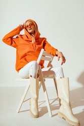 Fashionable woman wearing stylish orange hoodie, sunglasses, white skinny jeans, high boots sitting, posing on chair. Full-length studio portrait