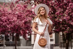 Fashionable elegant happy smiling woman wearing trendy spring, summer white dress, hat, with small shoulder round wicker bag, posing near blooming trees. Copy, empty space for text