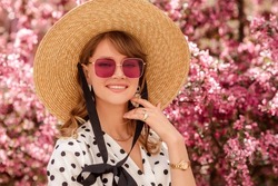 Elegant fashionable woman wearing trendy straw hat, pink square sunglasses, wrist watch, polka dot blouse, posing in street near pink spring blossom trees. Outdoor lifestyle portrait. Copy space 