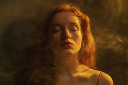 Beautiful redhead freckled woman posing in darkness, smoke and warm light. Art studio portrait. Copy, empty space for text
