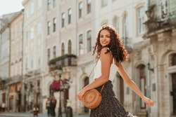 Happy smiling curly brunette woman wearing trendy summer outfit with round wicker shoulder rotan bag, white top, polka dot midi skirt, walking in street of European city. Fashion, lifestyle concept 