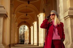 Fashionable confident blonde woman wearing trendy sunglasses, beret, pink turtleneck cashmere sweater, marsala color autumn coat, gloves, posing in street of European city. Copy, empty space for text