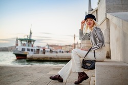 Elegant happy smiling fashionable woman wearing trendy baker boy cap, striped long sleeve shirt, white flared jeans, boots, holding small bag, posing near bridge in Venice. Copy, empty space for text