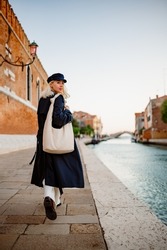 Elegant fashionable woman wearing trendy autumn navy blue trench coat, baker boy cap, with beige leather hobo bag, walking in street in Venice. Full-length outdoor portrait