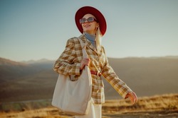 Happy smiling fashionable woman wearing stylish autumn outfit with orange hat, sunglasses, checkered blazer, belt, beige leather hobo bag, walking in mountain nature. Copy, empty space for text