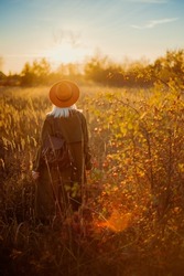 Rear, back view of fashionable woman wearing orange hat, olive green trench coat, with leather backpack, posing in beautiful autumn nature during the sunset. Copy, empty space for text