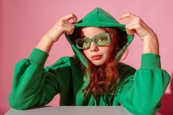 Fashionable freckled redhead girl wearing trendy green hoodie, stylish color rectangular glasses, posing against pink background. Close up studio portrait
