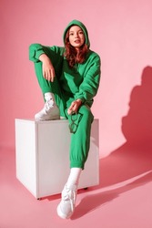 Happy smiling fashionable freckled redhead girl wearing trendy green suit with hoodie, joggers,  white sneakers, sitting, posing on pink background. Full-length studio portrait
