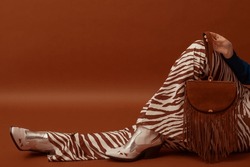 Trendy fashionable woman's outfit details: brown suede fringed bag, flared trousers with zebra print, white cowboy boots,. Copy, empty space for text