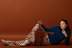 Fashionable confident woman wearing stylish blue turtleneck, trendy flared trousers with zebra print, white cowboy boots, holding brown suede fringed bag. Full-length studio portrait. Copy space
