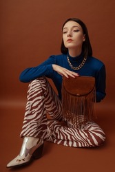 Fashionable confident woman wearing stylish blue turtleneck, trendy flared trousers with zebra print, white cowboy boots, holding brown suede fringed bag. Studio portrait 