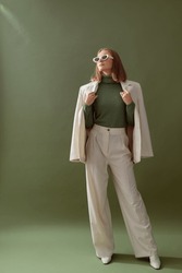 Fashionable confident woman wearing elegant white suit with blazer, wide leg trousers, cashmere turtleneck sweater, trendy sunglasses, posing on green background. Full-length studio fashion portrait