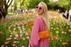 Happy smiling fashionable woman wearing trendy pink sunglasses, pleated dress, with orange faux patent leather baguette bag, posing among flowers. Copy, empty space for text