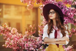 Outdoor portrait of young beautiful fashionable lady posing near flowering tree. Model wearing stylish accessories & clothes. Girl looking aside. Female beauty & fashion. City lifestyle. Copy space
