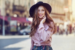 Outdoor portrait of young beautiful fashionable happy lady posing on a street of the old city. Model wearing stylish wide-brimmed hat & clothes. Girl looking at camera. Female fashion. City lifestyle