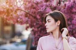 Outdoor close up portrait of  young beautiful girl posing on street, looking aside. Model wearing pink dress, has stylish earrings, hand watch. Female fashion concept. Copy, empty space for text