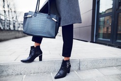 Fashionable woman posing in street, holding big dark blue textured bag, wearing stylish pointed toe ankle boots, elegant clothes. Female fashion concept. Copy, empty space for text.