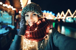 Outdoor close up portrait of young beautiful happy smiling girl making selfie photo in night street. Festive Christmas fair on background. Model looking at camera, wearing knitted beanie hat, scarf.