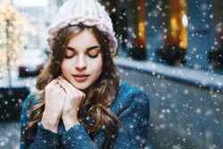 Outdoor close up portrait of young beautiful girl with long hair wearing hat, sweater posing in street of european city.  Christmas, winter holidays concept. Snowfall. Copy, empty space for text