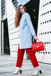 Outdoor full body portrait of young beautiful fashionable woman walking in street. Model wearing light blue coat, red pants, silvery ankle boots, holding trapeze handbag. Female fashion concept