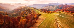 Autumn mountain panorama. Dirt road on top of the hills. Sunny landscape with meadow and colorful forest. Red, Yellow, Orange trees on hillsides. Carpathians, Ukraine, Europe.