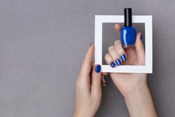 Female hands with manicure in trendy classic blue color holding white frame and a bottle of nail polish on the grey background