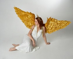 Full length portrait of beautiful red head woman wearing long flowing fantasy toga gown with golden halo crown and angel wings,  sitting pose isolated on a white studio background.