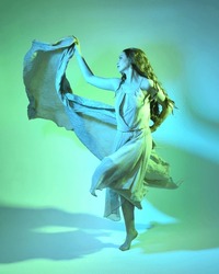 Full length  portrait of beautiful brunette woman dancer, wearing ethereal gown, dancing with flowing fabric.  isolated on green studio background with cinematic colourful lighting.