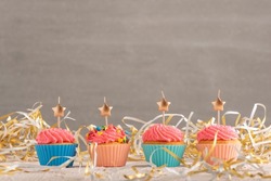 Golden candles in the shape of star on cupcakes. Muffins with pink buttercream frosting on festive tinsel background. Copy spacve on gray background.