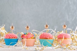 Golden candles in the shape of star on cupcakes. Muffins with pink buttercream frosting on festive tinsel background. Copy spacve on gray wall.